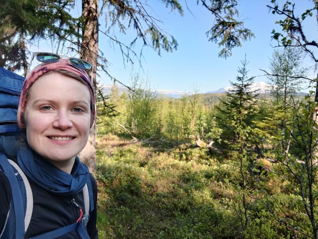 Ein Selfie einer Frau vor einer Landschaft mit Bäumen und Bergen im Hintergrund.