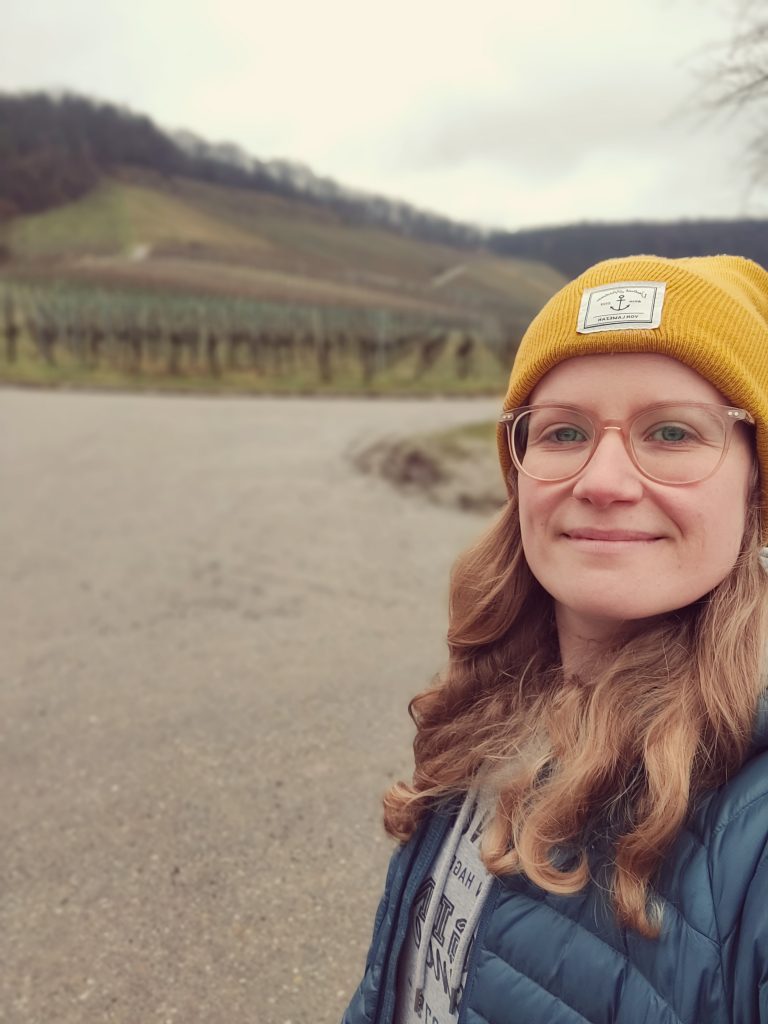 Ein Selfie einer Frau mit Mütze und Brille vor einem Weinberg.