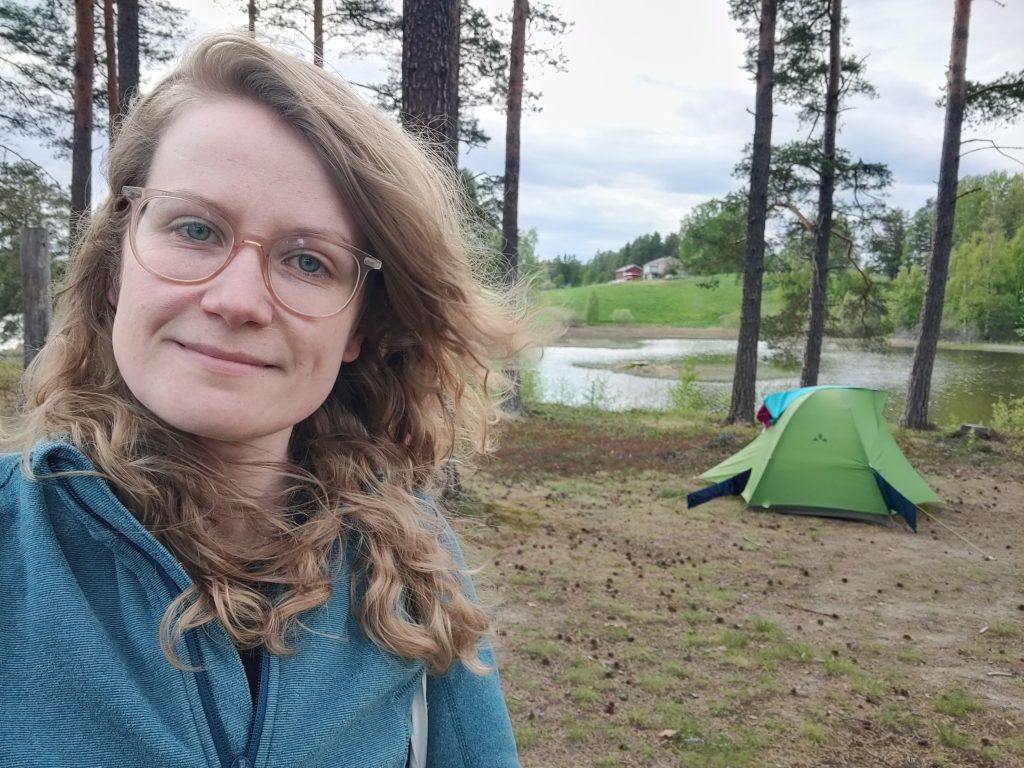 Ein Selfie einer Frau, die vor einer Lanschaft mit Bäumen, einem See und einem Zelt steht.