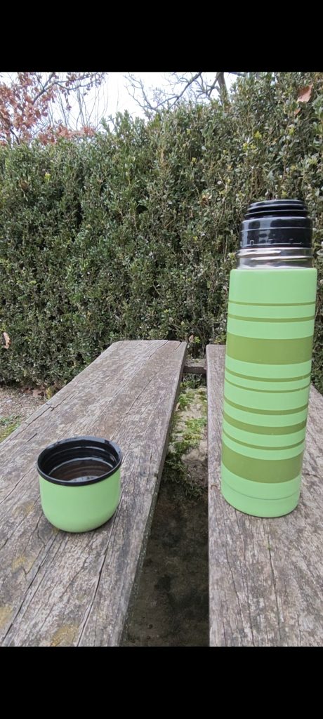 Eine Thermoskanne auf einer Bank. Im Hintergrund ist eine Hecke zu sehen.