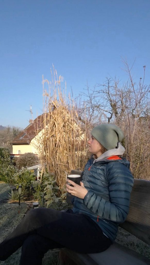 Eine Frau sitzt auf einer Bank und hat eine Tasse Kaffee in der Hand. Im Hintergrund ist ein Haus zu sehen.