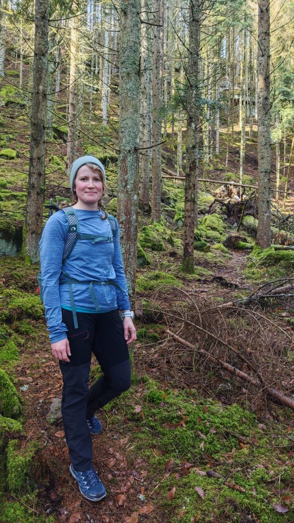 Eine Frau steht im Wald und trägt Outdoorkleidung.
